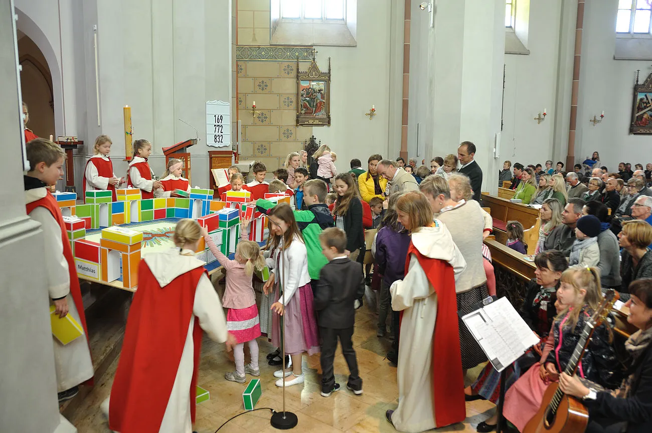 Familiengottesdienst: zahlreiche Kinder bauen mit den Ministrantinnen und dem Pfarrer im Altarraum ein himmlisches Jerusalem aus bunten Kartonziegeln. Auf manchen Ziegeln stehen die Namen der Apostel und die der getauften Kinder im vergangenen Jahr.